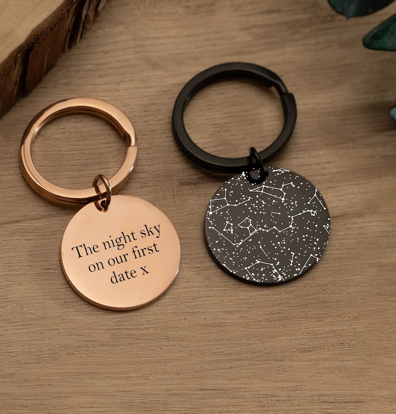 May include: Two keychains with round metal tags. One tag is rose gold with the text "The night sky on our first date x". The other tag is black with a white constellation design.