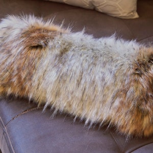 May include: A long, rectangular faux fur throw pillow with a brown, grey, and white colour scheme. The pillow is made of soft, fluffy fur and is perfect for adding a touch of luxury to any room.
