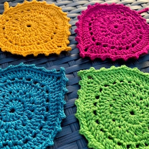 May include: Four crocheted coasters in different colors: yellow, pink, blue, and green. The coasters are made with a circular pattern and have a scalloped edge.