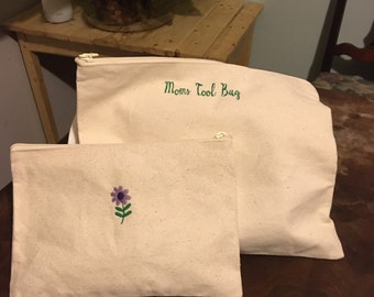 May include: Two canvas zipper pouches. The larger pouch has the text "Moms Tool Bag" in green. The smaller pouch features a purple flower with green stem embroidery. Both pouches are off-white.