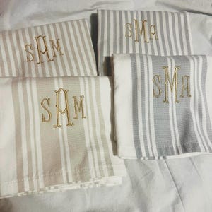 Puede incluir: Cuatro toallas plegadas con rayas verticales en gris, beige y blanco. Cada toalla presenta un monograma bordado en hilo dorado. Las toallas están dispuestas sobre una superficie blanca.