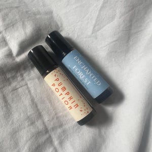 May include: Two roll-on bottles with black lids. One has a light beige label with "Pumpkin Potion" in orange text. The other has a light blue label with "Enchanted Forest" in white text. Both bottles are on a white surface.