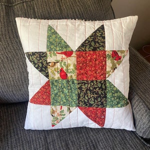 May include: A white cushion with a red, green and black star-shaped patchwork design featuring robins and holly. The cushion is quilted with white thread.