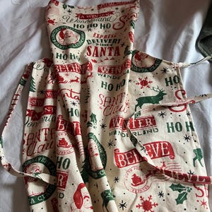 May include: Cream-colored Christmas apron with a festive print. The design features red and green Santa Claus imagery, reindeer, snowflakes, and the words "Santa," "Believe," and "Winter Wonderland." The apron has a neck loop and waist ties.