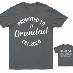 May include: A dark gray t-shirt with white text that reads "Promoted to Grandad Est. 2024" with a pair of white footprints above the word "Grandad". The back of the shirt has a small white text that reads "Name or small message on the back".