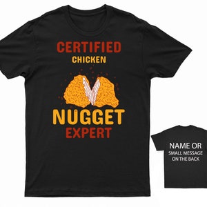 Op de afbeelding: Zwart T-shirt met de tekst "CERTIFIED CHICKEN NUGGET EXPERT" in oranje en rood. Een afbeelding van een kipnugget wordt in het midden van het shirt getoond.