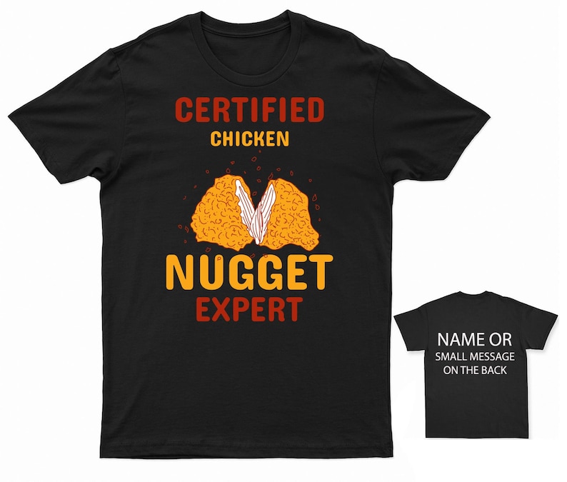 Op de afbeelding: Zwart T-shirt met de tekst "CERTIFIED CHICKEN NUGGET EXPERT" in oranje en rood. Een afbeelding van een kipnugget wordt in het midden van het shirt getoond.
