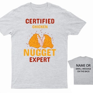 Op de afbeelding: Een grijs T-shirt met de tekst "CERTIFIED CHICKEN NUGGET EXPERT" in rood en oranje. Een cartoon afbeelding van een kippen nugget bevindt zich in het midden van het shirt. De achterkant van het shirt is grijs met de tekst "NAME OR SMALL MESSAGE ON THE BACK" in zwart.