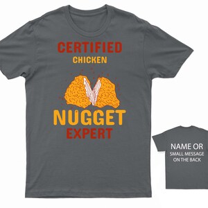 Op de afbeelding: Een donkergrijs T-shirt met de tekst "CERTIFIED CHICKEN NUGGET EXPERT" in rood en geel. Het shirt heeft een afbeelding van een kipnugget die doormidden is gebroken.
