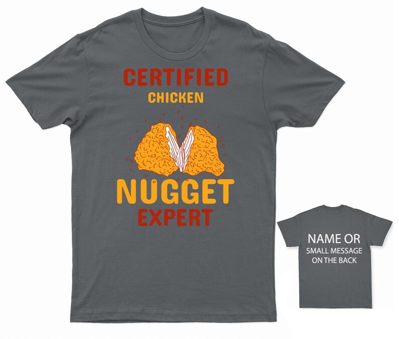 Op de afbeelding: Een donkergrijs T-shirt met de tekst "CERTIFIED CHICKEN NUGGET EXPERT" in rood en geel. Het shirt heeft een afbeelding van een kipnugget die doormidden is gebroken.