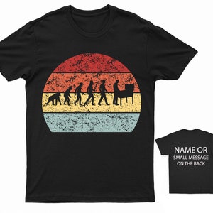 May include: Black t-shirt featuring a vintage-style graphic of the evolution of man culminating in a person playing a pinball machine. The graphic is set against a red, orange, yellow, and blue sunset.