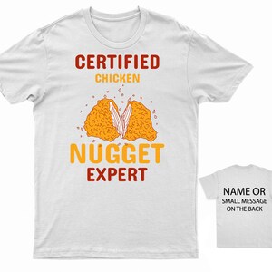 Op de afbeelding: Wit T-shirt met de tekst "CERTIFIED CHICKEN NUGGET EXPERT" in rood en oranje. Een cartoon afbeelding van een kipnugget bevindt zich in het midden van het shirt. De achterkant van het shirt is wit met de tekst "NAME OR SMALL MESSAGE ON THE BACK" in zwart.
