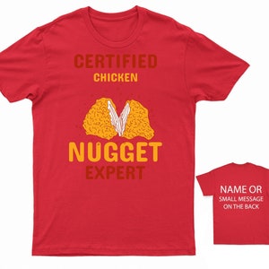 Op de afbeelding: Rood T-shirt met de tekst "Certified Chicken Nugget Expert" en een cartoon afbeelding van een kipnugget. De achterkant van het shirt heeft ruimte voor een naam of een klein bericht.