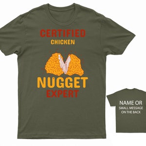 Op de afbeelding: Een groen T-shirt met de tekst "Certified Chicken Nugget Expert" in rood en geel. Het shirt heeft een afbeelding van een gehalveerd kippennugget.
