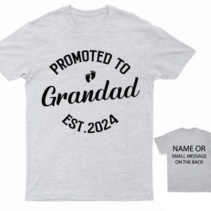May include: Gray t-shirt with black text that reads "Promoted to Grandad Est. 2024" with a pair of baby footprints above the word "Grandad".