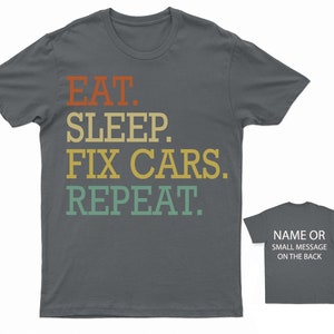 Puede incluir: Una camiseta gris oscuro con el texto "EAT. SLEEP. FIX CARS. REPEAT." en un esquema de colores retro. El texto est&aacute; en negrita, fuente sin serifas. La camiseta tiene cuello redondo y mangas cortas.
