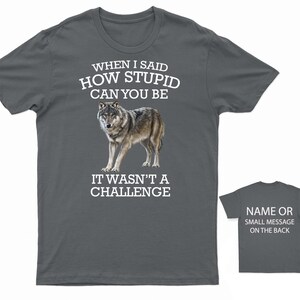 K&ouml;nnte beinhalten: Ein dunkelgraues T-Shirt mit einer wei&szlig;en Wolfgrafik und dem Text "WHEN I SAID HOW STUPID CAN YOU BE IT WASN'T A CHALLENGE". Auf der R&uuml;ckseite des Shirts steht "NAME OR SMALL MESSAGE ON THE BACK".