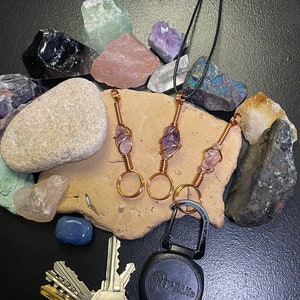May include: Three amethyst crystal pendants wrapped in copper wire, hanging from black cords. The pendants are displayed on a rock with other crystals and a black retractable key chain.