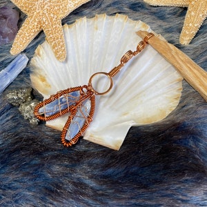 May include: A copper wire wrapped blue kyanite crystal pendant with a copper ring and chain. The pendant is displayed on a white seashell with a starfish and a piece of driftwood.