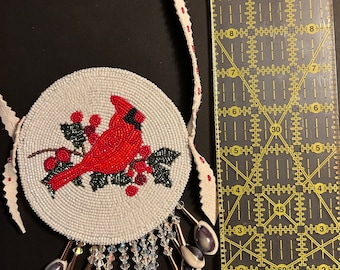 May include: A white beaded round ornament with a red cardinal bird and holly berries. The ornament is attached to a leather cord with a fringe of white and black beads.