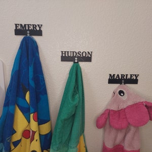 May include: Three towel hooks with personalized names, "Emery", "Hudson", and "Marley", are mounted on a white wall. A blue and yellow towel with a cartoon character is hanging on the "Emery" hook. A green towel is hanging on the "Hudson" hook. A pink towel with a cartoon animal face is hanging on the "Marley" hook.