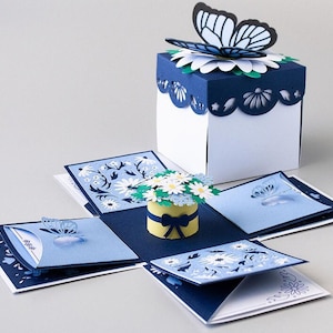May include: A blue and white papercraft explosion box with a butterfly on top. The box is decorated with flowers and butterflies. The box is open and the inside is visible. The inside of the box is decorated with flowers and butterflies.