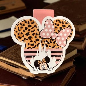 May include: A Minnie Mouse-themed bookmark featuring a leopard print design, pink polka dot bow, and a castle silhouette. The bookmark has a pink backing and is set against a backdrop of stacked books.