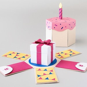 Puede incluir: Una caja de pastel de cumpleaños rosa y blanca con una sola vela encendida en la parte superior. La caja está abierta para revelar una caja de regalo blanca más pequeña con una cinta rosa y una base azul. La caja está rodeada de seis tarjetas de papel amarillas y azules con un diseño de bandera de fiesta.