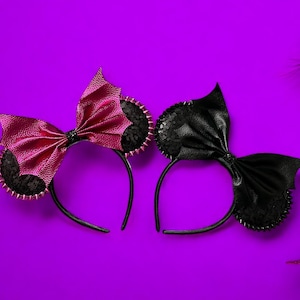 May include: Two black headband headbands with bows, one with a pink bow and one with a black bow. The headbands have black lace and silver studs. The headbands are on a purple background.