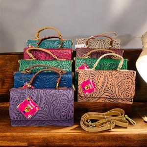 May include: A collection of nine colorful leather purses with intricate floral designs. Each purse has a top handle and a detachable shoulder strap. The purses are arranged in a stack on a wooden surface.
