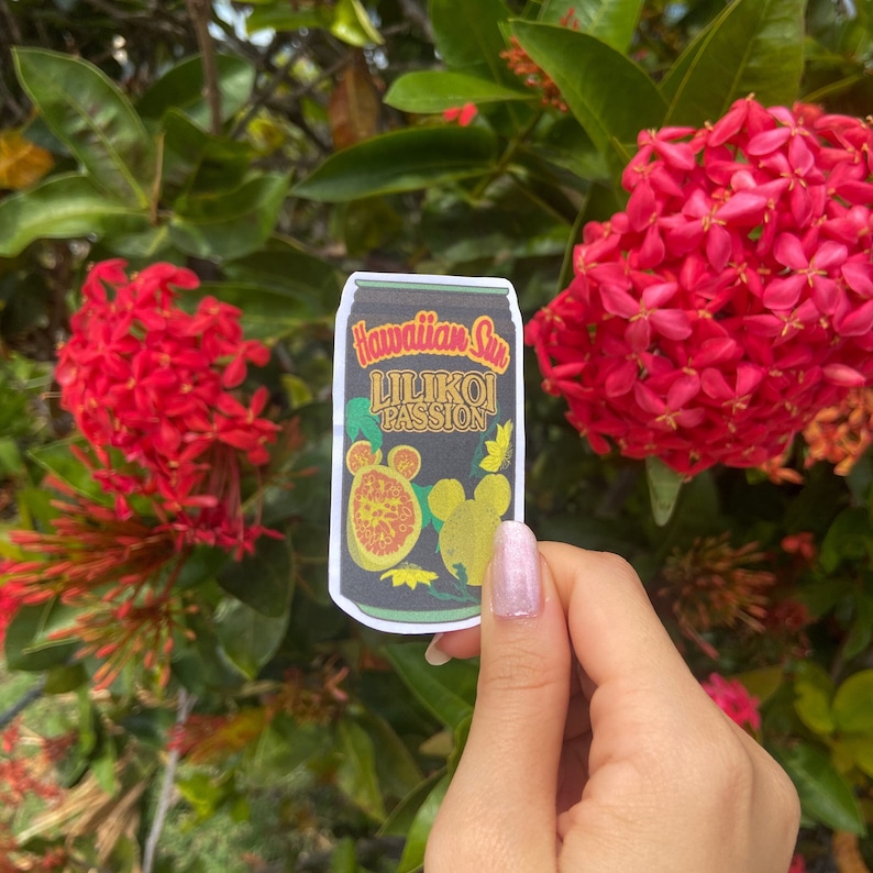 May include: A sticker featuring a black can design with the words "Hawaiian Sun Lilikoi Passion" in gold lettering. The sticker includes illustrations of passion fruit and a yellow butterfly. The background shows red flowers and green foliage.