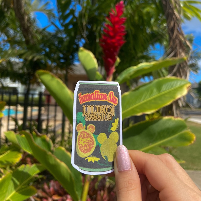 May include: A sticker featuring a black can design with the words "Hawaiian Sun Lilikoi Passion" in gold and green. The sticker includes illustrations of passion fruit and a yellow Mickey Mouse silhouette. The background shows green leaves and a red flower.