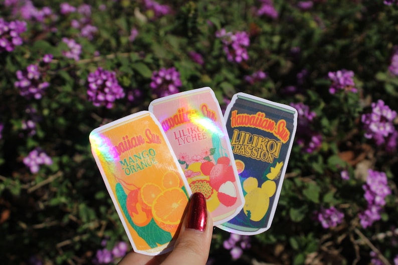 May include: Three stickers featuring Hawaiian Sun drink can designs. The stickers showcase Mango Orange, Lilikoi Lychee, and Lilikoi Passion flavors. The stickers have a holographic effect and are set against a background of purple flowers.