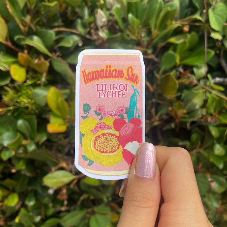 May include: A sticker featuring a pink can design with the words "Hawaiian Sun Lilikoi Lychee." The sticker showcases illustrations of passion fruit and lychee. The background is a lush green bush.