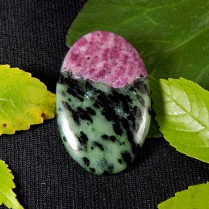 May include: An oval-shaped polished gemstone featuring a distinct color split. The top section is a vibrant magenta pink, while the bottom portion is a pale mint green speckled with irregular black inclusions throughout the lower surface area.
