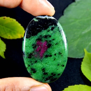 Könnte beinhalten: Ein oval geschliffener Edelstein mit einer leuchtend grünen Grundfarbe, durchsetzt mit markanten schwarzen Sprenkeln und einem deutlich abgegrenzten, konzentrierten Fleck in tiefem Magentarosa in der Mitte der glatten, reflektierenden Oberfläche.