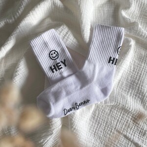 Könnte beinhalten: Ein Paar weiße Knöchelsocken mit schwarzem Text. Eine Socke sagt "HEY" mit einem Smiley und die andere sagt "HI". Die Socken haben den Text "Dear Benno" auf der Unterseite.