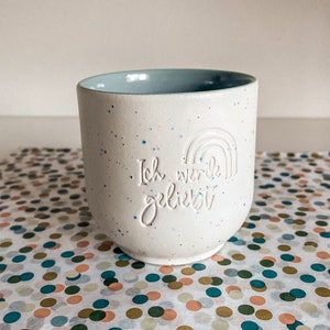 Könnte beinhalten: Weiße Keramiktasse mit hellblauem Rand und Farbsprenkeln. Die Tasse hat ein Regenbogen-Design und den Text "Ich werde geliebt" in Weiß.