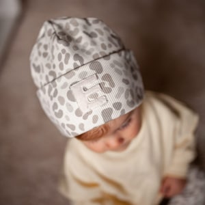 Könnte beinhalten: Ein Baby trägt eine weiße und graue Mütze mit Leopardenmuster und einem weißen Buchstaben "E" auf der Vorderseite. Das Baby trägt einen cremefarbenen Pullover und schaut nach unten.