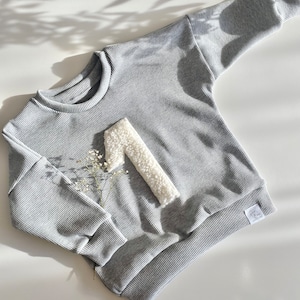 Könnte beinhalten: Ein grauer gerippter Pullover mit einer weißen, flauschigen Zahl eins auf der Vorderseite. Der Pullover hat ein weißes Etikett mit dem Text "Joy & Bloom" darauf.