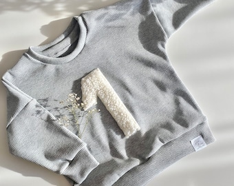 May include: A gray ribbed sweatshirt with a white fuzzy number one on the front. The sweatshirt has a white tag with the text "Joy & Bloom" on it.