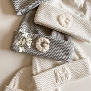 Könnte beinhalten: Vier gestrickte Mützen in Grau und Weiß mit flauschigen weißen Buchstaben, die darauf genäht sind. Die Buchstaben sind J, G, M und M.