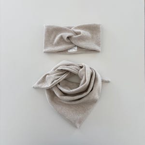 Könnte beinhalten: Beiges geripptes Strick-Stirnband und Schal-Set. Das Stirnband hat ein verdrehtes Knotendesign. Der Schal ist dreieckig und geknotet. Beide Artikel sind aus einem weichen, neutralfarbenen Stoff gefertigt, ideal für einen abgestimmten Look.