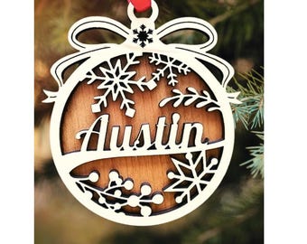 Peut inclure: Ornement de Noël en bois avec le nom "Austin" découpé. L'ornement présente un nœud, des flocons de neige et un ruban rouge pour l'accrocher. L'arrière-plan est une image floue d'un arbre de Noël.