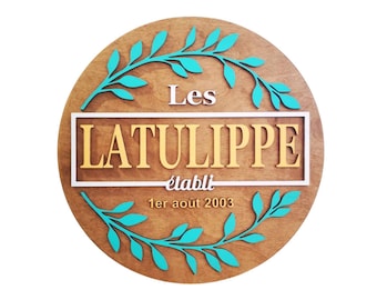 May include: A round wooden sign with a white frame and the text "Les LATULIPPE établi 1er août 2003". The sign is decorated with a teal green leafy wreath.