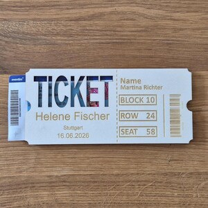 Könnte beinhalten: Ein weißes Ticket mit dem Wort "TICKET" ausgeschnitten, das ein Konzertbild zeigt. Das Ticket ist für Helene Fischer in Stuttgart am 16. Juni 2026, mit Details wie Block 10, Reihe 24 und Sitz 58.