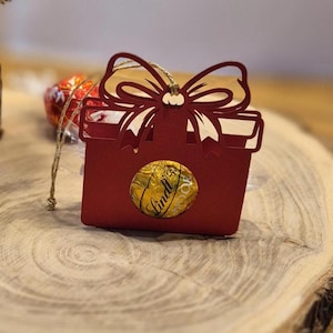 Könnte beinhalten: Eine rote Geschenkbox mit einem dekorativen Schleifenausschnitt oben. Auf der Vorderseite der Box ist eine goldene Lindt-Schokoladenkugel zu sehen. Die Box steht auf einer Holzoberfläche.