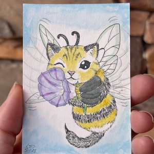 May include: A watercolour illustration of a cat-bee hybrid holding a purple flower. The artwork features a yellow and black striped body, transparent wings, and a winking expression. The background is light blue.