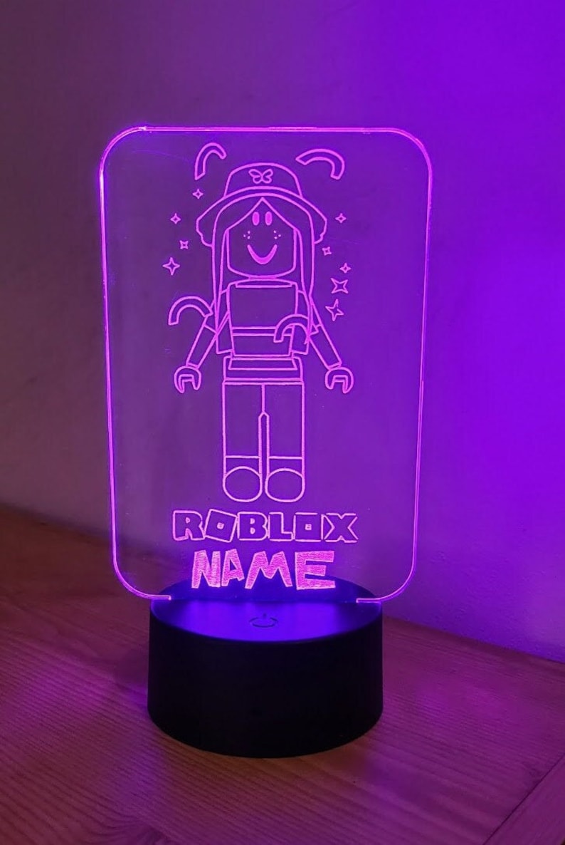 May include: A purple light-up nightlight with a clear acrylic panel featuring a line drawing of a Roblox avatar wearing a hat and a bow. The text "ROBLOX NAME" is below the avatar.