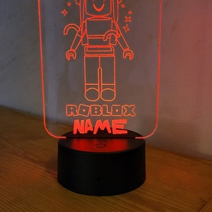 May include: A red acrylic night light with a Roblox character design. The light features a female Roblox avatar wearing a hat and holding a butterfly. The text "ROBLOX NAME" is printed below the character.